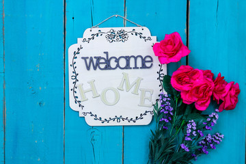 Welcome Home sign with purple and red flowers hanging on rustic antique teal blue wood background