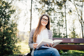 young woman sitting on bench in park, reading book, smiling