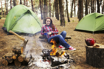 Camping in the forest. The girl sits in a hammock and looks at the fire.