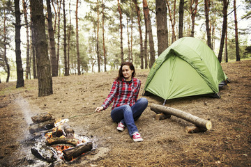 Camping in the forest. The girl sits in a hammock and looks at the fire.