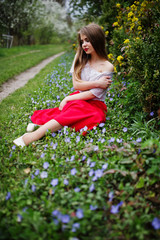 Portrait of sitiing beautiful girl with red lips at spring blossom garden on grass with flowers, wear on red dress and white blouse.