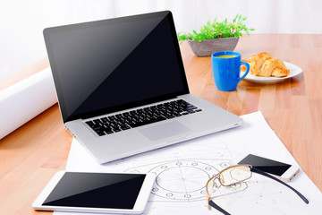 Workplace of cad specialist with laptop, smartphone, tablet, glasses, blue cup and draws on wooden table.