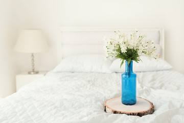Fototapeta na wymiar White flowers in blue glass bottle flower pot on wooden serving board on the bed in white room