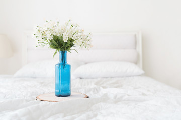 Flowers in blue bottle flower pot on bed. White bedroom interior details
