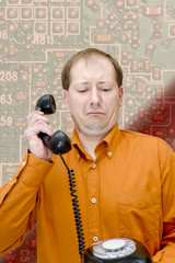 A white man in an orange shirt with a telephone receiver in his hand looks at the old black disk-wired telephone in amazement and distrust with a background of a board with electronic components.