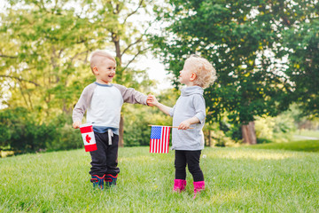 Happy adorable little blond Caucasian girl and boy smiling laughing holding hands and waving American and Canadian flags, outside in park, celebrating 4th july Independence Day
