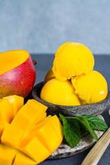 Mango ice cream sorbet with mint leaves and mango fruit in black stone bowl on grey textured...