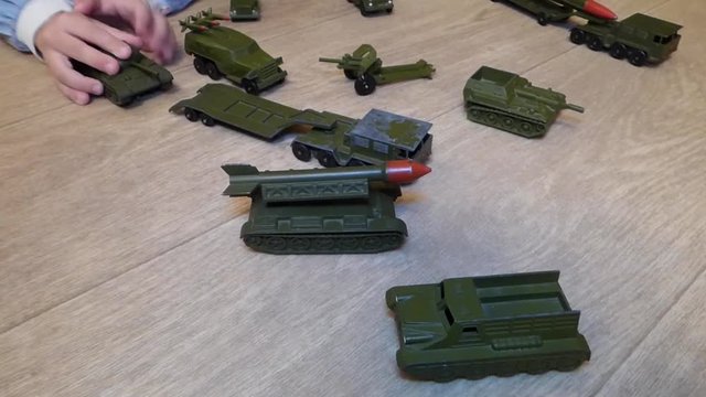 Kid rolls old military vehicles - metal cars colored in khaki. Soviet (russian) retro toys: tanks, trucks, missiles and cannon. Caucasian child plays war. Little army on the floor.