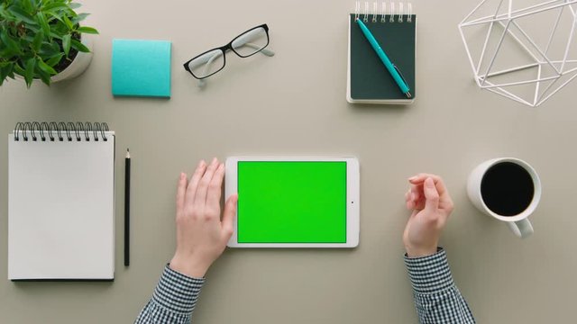 Top view woman using tablet device with green screen at office. Table background with white and blue objects, items. Hands scrolling, swiping pages. Chroma key.