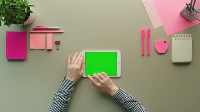 Woman using white tablet device with green screen on grey table background with pink objects. Female hands tapping, swiping, scrolling pages on laptop computer. chroma key