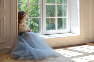 Anonymous shoot of cute five years blonde girl in blue fluffy gorgeous dress with bouquet of pink hydrangea flowers sitting back to the camera opposite the large window with green garden lush outside