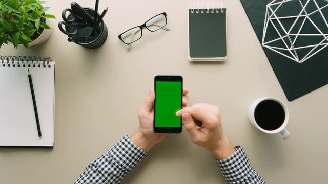 Top view man holding smartphone with green screen at office table background. Man hands tapping on mobile phone touchscreen, scrolling, swiping pages. Chroma key