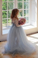 Anonymous shoot of cute five years blonde girl in blue fluffy gorgeous dress with bouquet of pink hydrangea flowers standing back to the camera opposite the large window with green garden lush outside