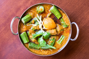 Vegetarian okra curry dish with fresh veggies, served in traditional steel bowl on mahogany table. Top view. - obrazy, fototapety, plakaty