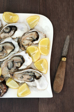 Oysters on ice on a porcelain plate with lemon fruit and antique oyster knife on old oak wood background.
