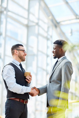 Portrait of smiling businessman greeting African-American partner by handshake standing in modern...
