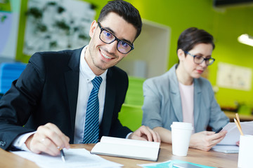 Happy young man looking at camera by workplace while reading papers