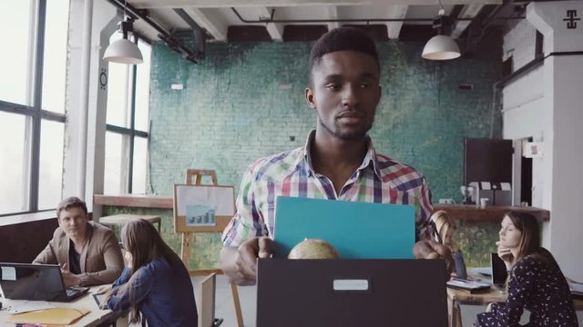 Young african man get fired from work at start-up. Male walks through the office, carrying box with personal belongings.