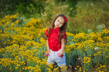 Young cute girl in red shirt and white shorts standing close to yellow flowers.