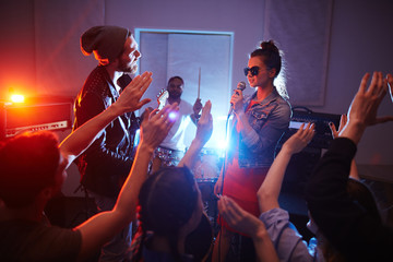 Cool band singing on stage of urban club with crowd cheering and dancing people partying, raising...