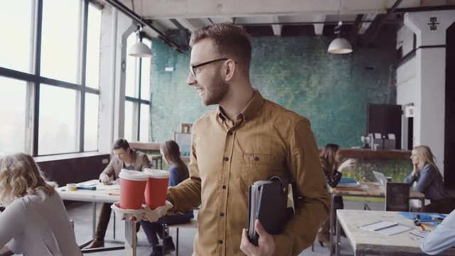 Handsome businessman manager comes to modern office at work. Young male greets with colleagues, brings coffee to friend.