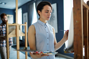 Portrait of young beautiful woman painting picture on easel standing in row of students in art class looking focused and concentrated