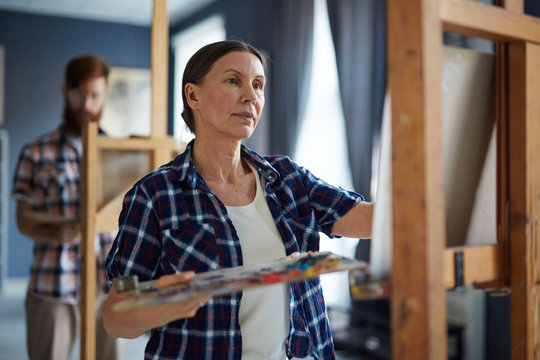 Portrait Of Elegant Mature Woman Painting Picture On Easel Standing In Row Of Students In Art Class