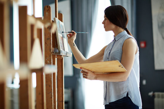 Portrait of talented young woman painting picture on canvas in art studio with inspiration