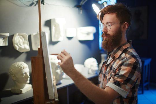 Portrait of inspired young red-haired man drawing plaster heads on canvas in art studio, looking focused and concentrated