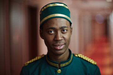 Portrait of smiling young bellhop of African ethnicity working in hotel, welcoming tourists looking at camera - obrazy, fototapety, plakaty