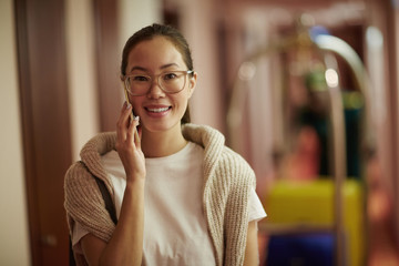 Portrait of smiling Asian woman standing in hotel hall with bags, speaking by smartphone and looking at camera