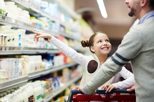 Happy family buying groceries: smiling little girl choosing dairy products from fridge in milk aisle while shopping in supermarket