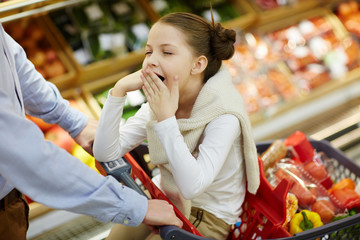 Portrait of little tired girl yawning sitting in shopping cart while parents buy groceries
