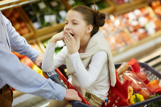 Portrait of little tired girl yawning sitting in shopping cart while parents buy groceries