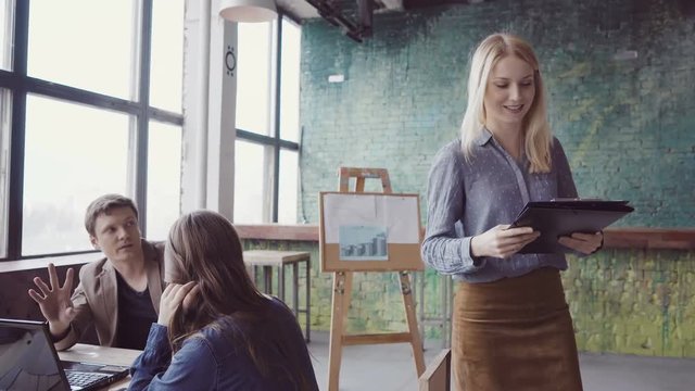 Young blonde team leader walking through modern office and controls work of employee. Female looks the fabric examples.