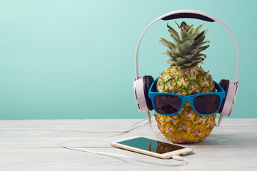 Pineapple with sunglasses, headphones and smart phone on wooden table over mint background. Tropical summer vacation and beach party concept.