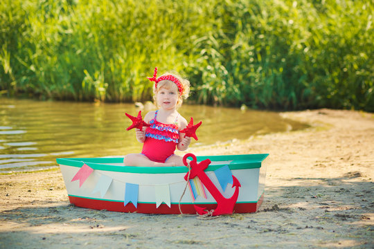 A cute little girl in a red bathing suit standing on the beach against the backdrop of boats