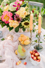 picnic, food, summer, holiday concept - close up on part of the festively decorated table with a beautiful bouquet of roses and peonies, candles in candlesticks, apricots, cherries, pear, glassware