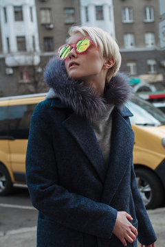 A blonde girl with glasses and a coat walks the streets of the city