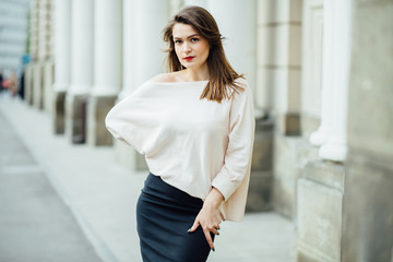 Slim elegant woman with red lips in a beige blouse and black skirt on the white building background.