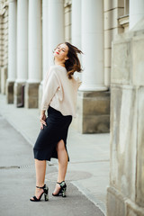 Obraz premium Slim elegant woman with red lips in a beige blouse and black skirt on the white building background. Vertical framming, full body portrait.