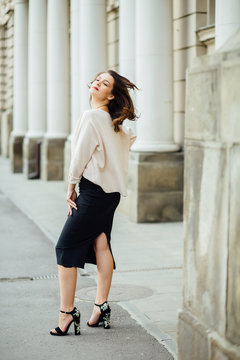 Slim elegant woman with red lips in a beige blouse and black skirt on the white building background. Vertical framming, full body portrait.