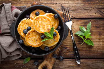 Delicious cottage cheese pancakes or syrniki with fresh blueberry in cast-iron pan on dark wooden rustic background, above view. Tasty breakfast.