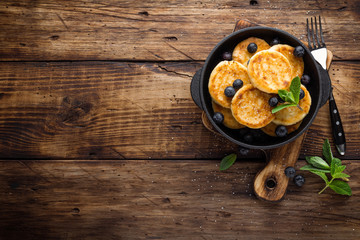 Delicious cottage cheese pancakes or syrniki with fresh blueberry in cast-iron pan on dark wooden rustic background, above view. Tasty breakfast.