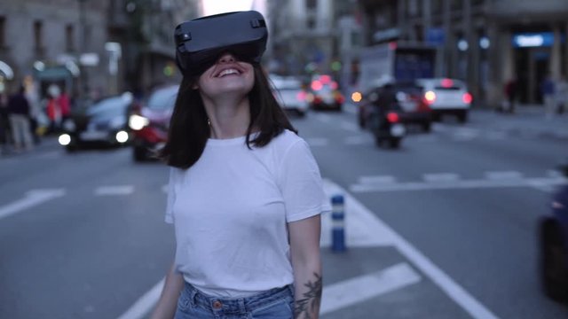 Excited laughing and smiling young brunette girl has unforgettable emotional experience using vr headset on city streets busy with traffic at evening