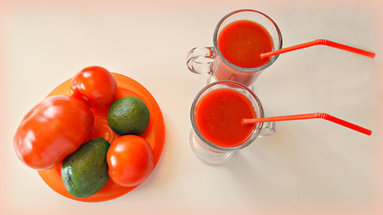  А number of tomatoes, avocado and 2 cups of tomato juice with straws on a white table. 