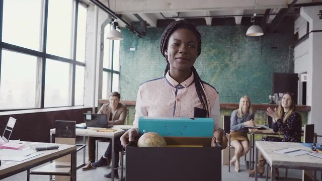 Young african woman recently hired for corporate job comes into new office. Female holds box with personal belongings.