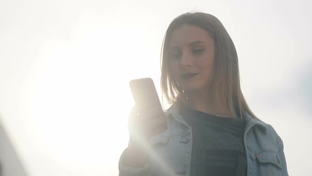 Satfisfied smiling cute youthful girl similar to famous actress with dark-red lips in blue denim court and strict bluse on shining sun and using her phablet cellphone on the city urban background