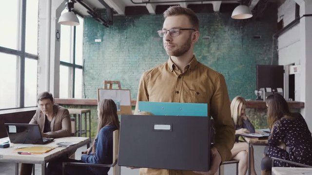 Handsome young man getting fired from work. Sad male walks through the office, carrying box with personal belongings.