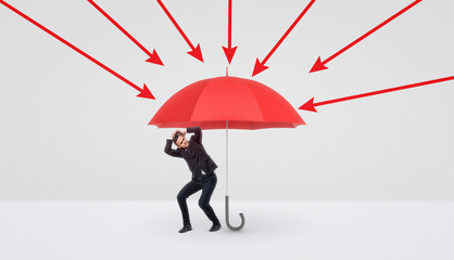 A small businessman hiding under a large red umbrella that protects him from red arrows.